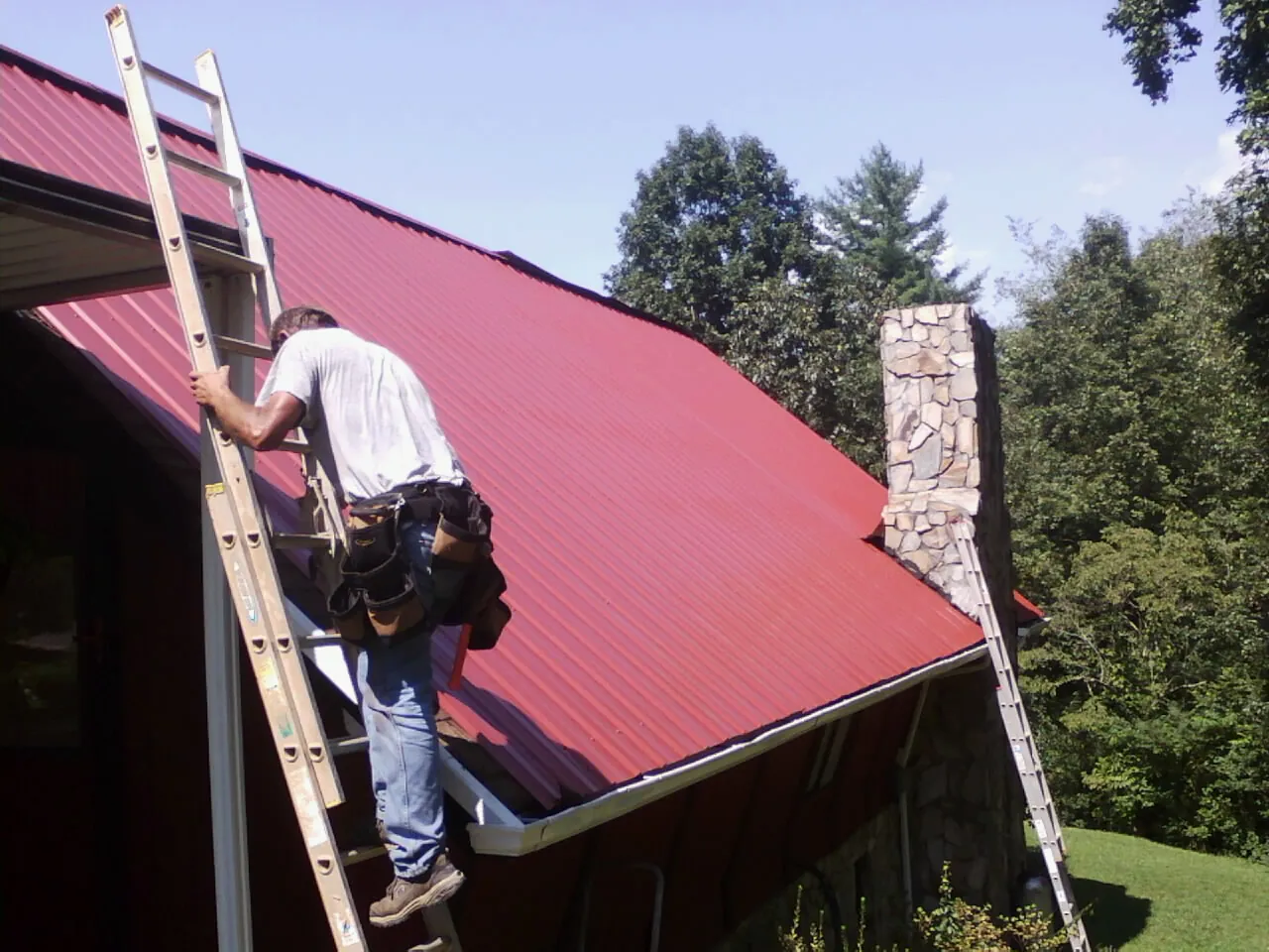 Roofer on a ladder beside a red metal roof and stone chimney — Soffit & Fascia Repair by Roofing Brothers in Manasquan